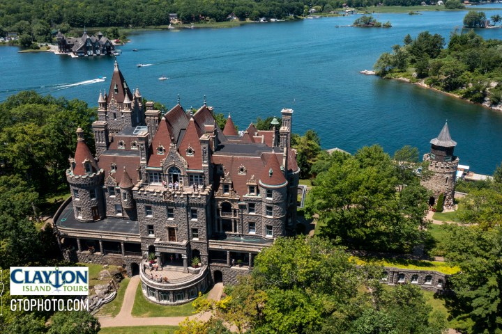 a castle on top of a body of water surrounded by trees with Thousand Islands in the background