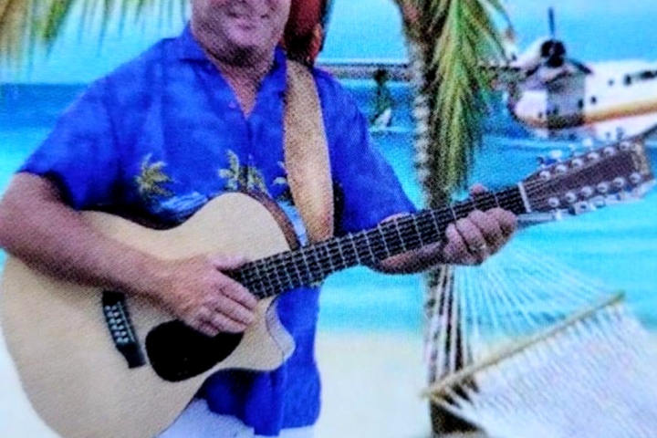 Man in tropical shirt with guitar and parrot on shoulder, beach backdrop with seaplane.
