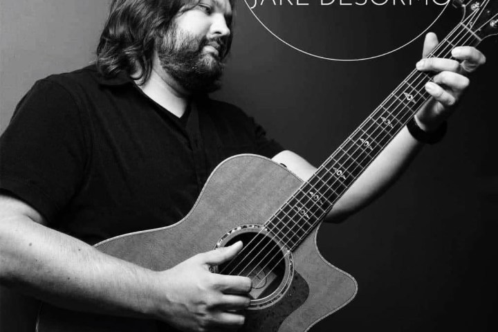Man playing acoustic guitar, logo 'JD Jake Desormo' beside him, grayscale image.