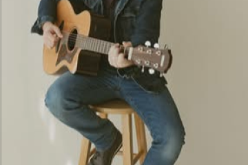 Man sitting on stool playing acoustic guitar, wearing denim outfit.